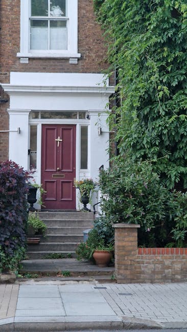 The image depicts the entrance to a period brick townhouse with a deep red front door, surrounded by white decorative architectural trim, situated at the top of a short flight of concrete steps. To th